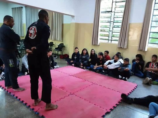 Professores de quimono preto conduzindo atividade com estudantes sentados ao redor do tatame em uma escola