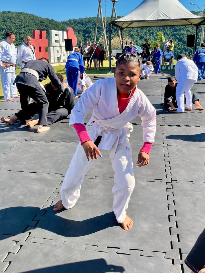 Aluna de quimono branco em posição de luta no tatame, com outros alunos treinando ao fundo