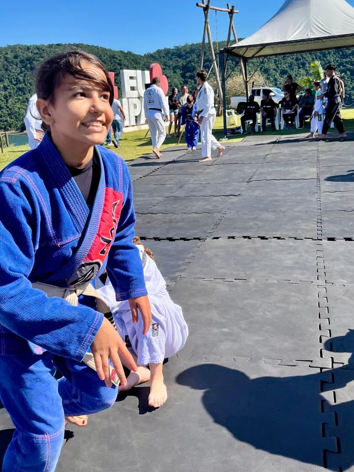 Aluno de quimono azul sorrindo, agachado no tatame durante aula ao ar livre